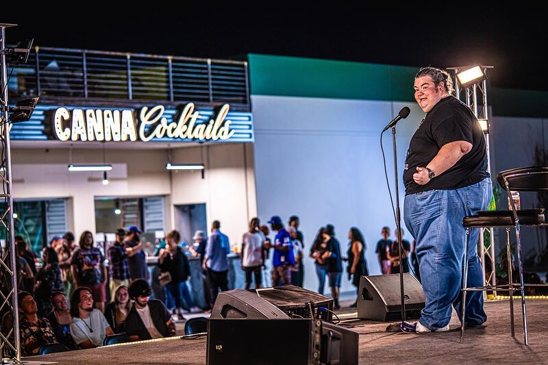 A comedian on the NuWu courtyard stage at night, lit by the CANNA Cocktails neon